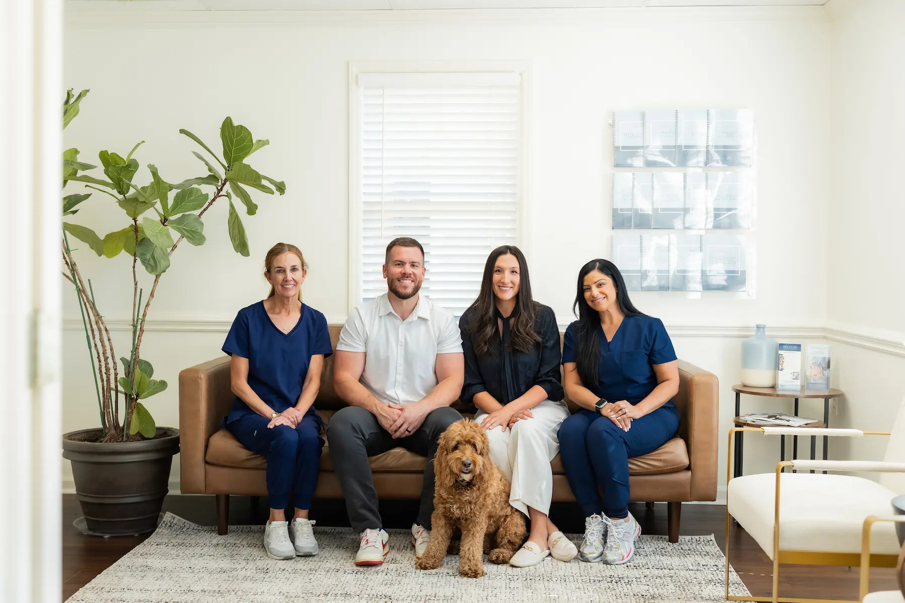 Four people and a golden-brown dog sit on a brown sofa in a bright room. Two women in blue scrubs, a man in a white shirt, and a woman in a black blouse smile at the camera.